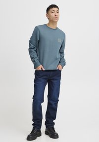 Felpa azzurra chiara con scollo rotondo e dettaglio logo dorato, abbinata a jeans blu scuro e stivali neri con lacci. Tessuto morbido.