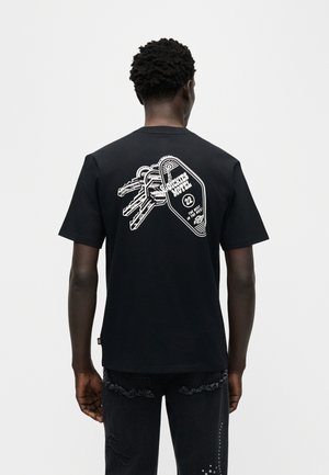 Άνδρας που φοράει μαύρο μπλουζάκι με λευκό σχέδιο κλειδιών και μπρελόκ μοτέλ εκτυπωμένο στην πλάτη, στέκεται μπροστά σε απλό φόντο.