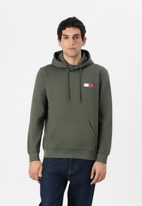 Felpa color verde oliva con tasca a marsupio, cappuccio con lacci e un piccolo logo sul petto con i colori rosso, bianco e blu. Tessuto morbido al tatto.