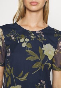 Blusa azul-marinho com design floral, apresentando flores em verde claro e rosa. Mangas curtas e transparentes com decote redondo. Textura suave.