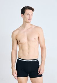Schwarze Boxershorts mit einem weißen "HUGO"-Logo am Bund, aus einem glatten, elastischen Stoff, der eng am Körper anliegt.