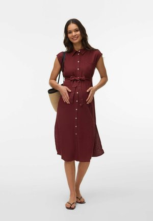 Lachende vrouw met lang donker haar, gekleed in een mouwloze bordeauxrode jurk met knopen, zwarte sandalen, met een strooien shopper, staand met haar handen in haar zij.