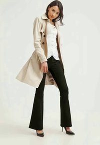 Casaco trench bege com botões, sobre uma blusa de renda branca e calças justas pretas. Saltos altos completam o conjunto, enfatizando um visual estruturado.