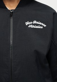 Μαύρο αθλητικό μπουφάν με φερμουάρ και λευκό λογότυπο "New Balance Athletics" στο αριστερό στήθος, φορεμένο πάνω από λευκό πουκάμισο.