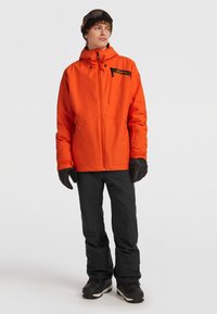 Oranje waterdichte jas met capuchon en zwarte accenten, gecombineerd met zwarte broek en handschoenen; geschikt voor wintersport.