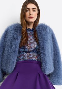 Faux fur jack in lichtblauw, transparante bloemtop met donkerblauwe en paarse accenten, en een effen paarse rok met plooien.