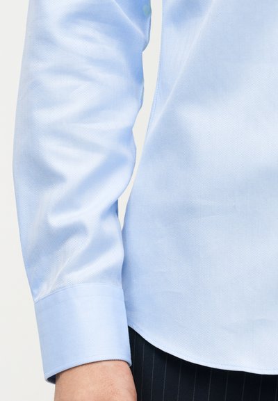 Μανσέτα και κάτω μανίκι μακρυμάνικου πουκάμισου ανοιχτού γαλάζιου με διακριτικό σχέδιο ψαροκόκαλου, φορεμένο με σκουρόχρωμο παντελόνι με λεπτές κάθετες ρίγες.