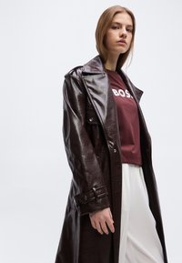 Νέα γυναίκα που φοράει σκούρο καφέ δερμάτινο παλτό τύπου trench, μπορντό μπλουζάκι BOSS και λευκό παντελόνι, ποζάροντας μπροστά σε απλό φωτεινό φόντο.