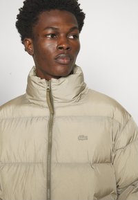 Chaqueta acolchada beige con cuello alto, cierre de cremallera y tela texturizada. Presenta un pequeño logotipo bordado en el pecho.