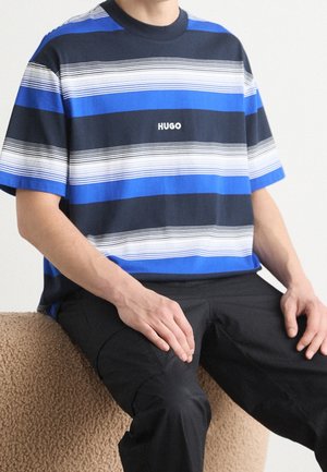 Person sitzt und trägt ein blau-schwarz-weiß gestreiftes T-Shirt mit dem Schriftzug "HUGO" und schwarze Hose, Hände ruhen auf den Oberschenkeln.