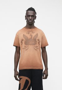 Jaunas tamsios odos vyras su trumpais dreideliais, dėvintis suplonintą oranžinę grafinę marškinėlę ir juodus šortus su dideliu ruduo logotipu.