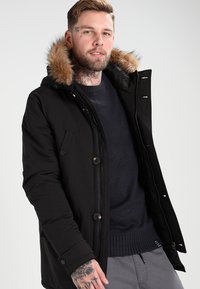 Svart parkajakke med pelsfôret hette, trykknapper og glidelås foran, kombinert med en marineblå genser under. Strukturert stoff.