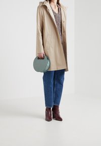 Beige wasserdichter Mantel mit Kapuze, mehrfarbiger Strickpullover, blaue Jeans, burgunderrote Stiefeletten und eine grüne runde Handtasche mit Henkel.