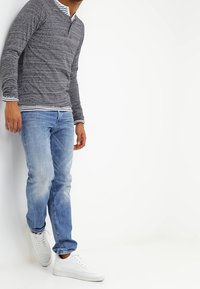 Man som bär en grå långärmad skjorta, över en randig skjorta, blå jeans och vita sneakers, står mot en vit vägg.