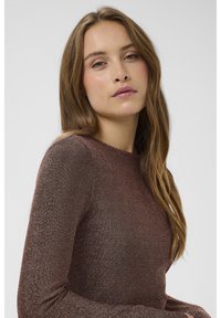 Ruskea metallinhohtoinen pitkähiha paita, jossa on sileä pinta, pyöreä pääntie ja vartalonmyötäinen malli, pukeutuneena aaltoilevat hiukset omaavaan malliin.