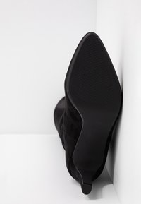 Botte à talon haut en suède noir à bout pointu, positionnée en angle dans un coin blanc, montrant la semelle texturée et un talon bob.