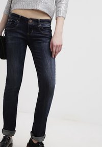 Jeans de denim azul oscuro con corte ajustado, que presentan un diseño clásico de cinco bolsillos, puños enrollados y una sutil textura desgastada.
