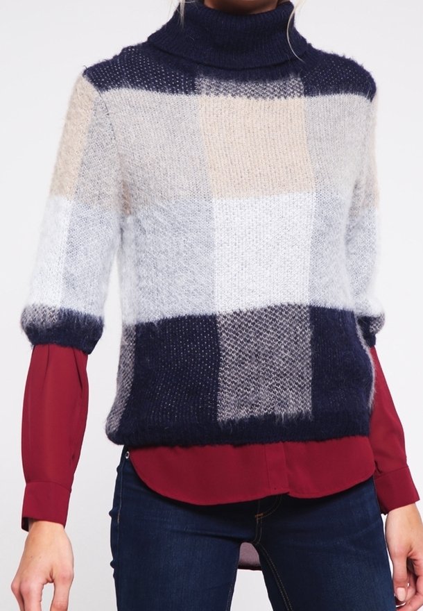 Kvinna i en flerfärgad rutiga turtleneck-tröja över en röd långärmad skjorta och mörkblå jeans.