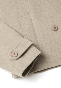 Detalle de abrigo de lana beige con una manga doblada, que presenta una textura suave y un botón marrón con cuatro orificios. Diseño simple y limpio.