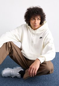 Balta sweatshirt su juodu Nike logotipu, derinama su rudomis kelnėmis ir pilkais sportbačiais. Medžiaga minkšta, o dizainas pasižymi laisvu siluetu.