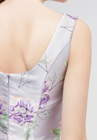 Kukallinen mekko vaaleanharmaasta kankaasta, jossa on violetteja ja vihreitä kukkakuvioita. Mekossa on avara selkäosa ja piilotettu vetoketjusuljenta.