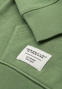 Sudadera verde con cuello acanalado y bolsillo. Presenta una etiqueta cosida con el texto "TU ÚNICO LÍMITE ES TU MENTE" y detalles de la marca.