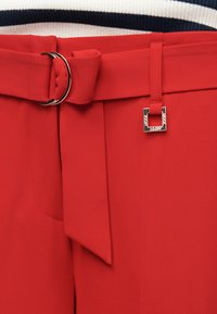 Pantalon rouge avec une ceinture en tissu assorti attachée à travers une boucle double anneau argentée et un petit détail décoratif carré argenté.