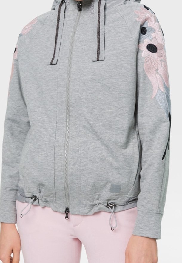 Sweat à capuche zippé gris clair avec cordons et broderie florale rose sur les épaules, porté avec un pantalon rose clair.