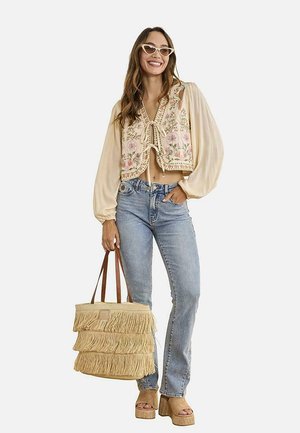 Mujer sonriente, con blusa crema bordada con flores y mangas abullonadas, vaqueros de lavado claro, sandalias de plataforma, gafas de sol de ojo de gato, sosteniendo un bolso tote con flecos.