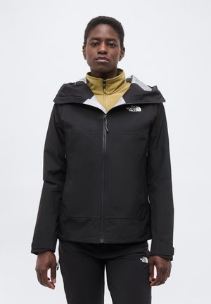 Άτομο που φοράει μαύρο μπουφάν με φερμουάρ και κουκούλα και μαύρο παντελόνι, με μπεζ εσωτερικό στρώμα ορατό στον λαιμό, στέκεται μπροστά σε λευκό φόντο.