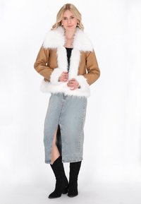 Frau in einer beigefarbenen Jacke mit weißem Kunstfellbesatz, hellblauer Jeansrock mit vorderem Schlitz, schwarzen Stiefeln und Ohrringen, die vor einem weißen Hintergrund steht.