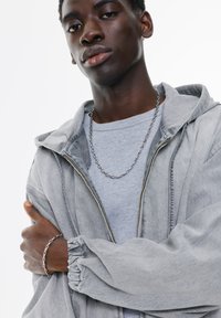 Siva zip-up kaput s kapuljačom s nabranim rukavima, nošen preko svijetlosive rebraste majice. Sadrži srebrni lanac kao ogrlicu i srebrnu narukvicu.
