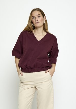 Pikade blondide juustega naine, kes kannab lahtist burgundia V-kaelusega pluusi ja beeže pükse, seisab valgel taustal, käed taskutes.