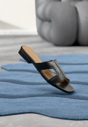 Sandalo nero senza lacci con design intagliato, posizionato su un tappeto blu testurizzato con un oggetto grigio morbido e curvo sullo sfondo.