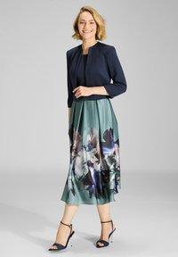 Chaqueta recortada navy sobre una blusa oscura y una falda teal con estampado floral, con tela fluida y sandalias de tiras con tacón delgado.