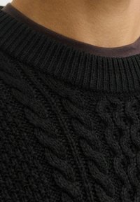 Close-up van de kraag van een zwart kabelgebreid sweater, gedragen over een bruine crewneck tee, waarbij de gedetailleerde gebreide textuur en de geribbelde halslijn goed zichtbaar zijn.
