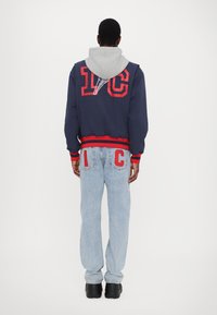 Marine bomberjakke med røde detaljer og grå hætte, parret med lyseblå jeans med røde "I" og "C" på baglommerne, sorte sko.