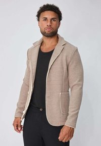 Homme aux cheveux bouclés portant un blazer texturé beige sur une chemise noire et un pantalon noir, se tenant contre un fond clair.