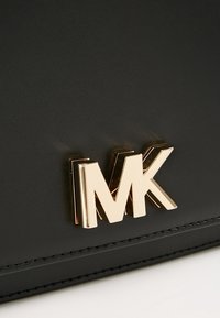 Logo métallique doré "MK" sur une surface en cuir noir lisse avec des coutures visibles le long du bord.