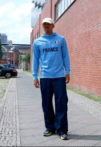 Hellblaues Zip-Hoodie mit "FRANCE" in fettem schwarzen Schriftzug und einer kleinen Flagge, kombiniert mit lockeren dunkelblauen Jeans und schwarzen Sneakers.