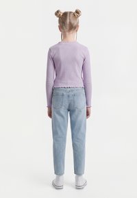 Mergina su šviesiais dvigubais kuodeliais, dėvinti levandų spalvos ilgomis rankovėmis marškinėlį, šviesiai mėlynas džinsus, baltas kojines ir baltus sportinius batelius, stovinti atsukus nugarą.