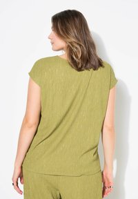 Top verde texturizado con mangas cortas, que presenta un ajuste holgado y sutiles acentos de hilo metálico. Vista trasera, sin patrones ni detalles adicionales.