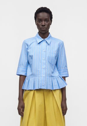 Femme portant une chemise bleue rayée boutonnée avec des manches jusqu'au coude et une jupe jaune plissée, debout devant un fond uni.