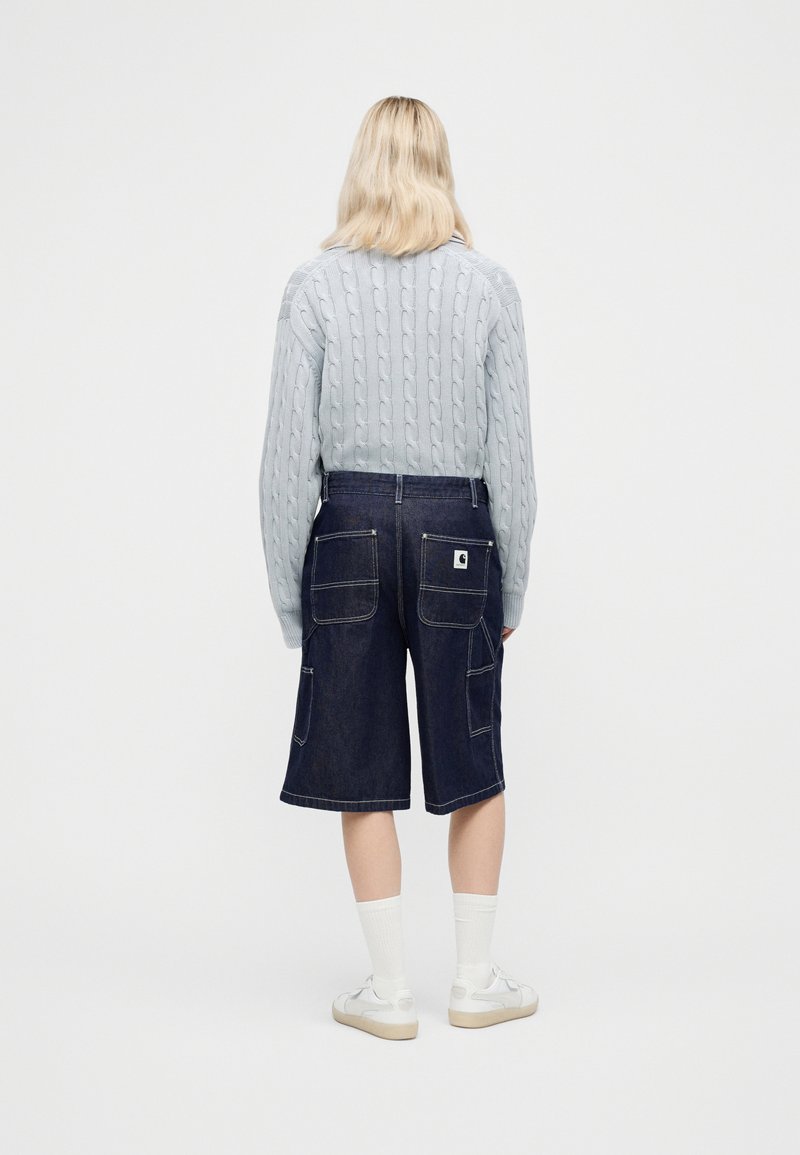 Personne aux cheveux blonds portant un pull en maille torsadée bleu clair, un short en denim foncé arrivant aux genoux avec de grandes poches, des chaussettes blanches et des baskets blanches.