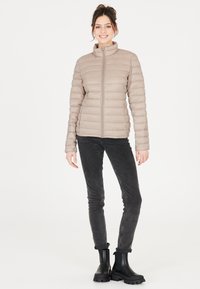 Lichtbeige pufferjas met een horizontaal doorgestikt patroon, hoge kraag en een rits aan de voorkant. Gepaard met zwarte slim-fit jeans en stevige zwarte laarzen.