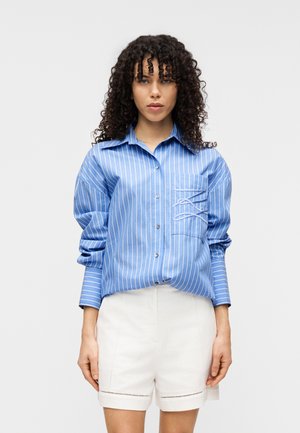 Blau-weiß gestreiftes Hemd mit Knöpfen und einer Tasche; zeichnet sich durch eine lockere Passform und hochgekrempelte Ärmel aus, kombiniert mit weißen Shorts.
