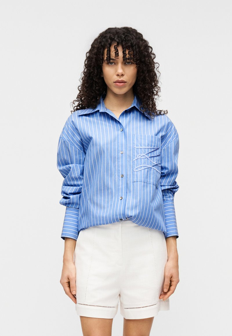 Μπλε και λευκό ριγέ πουκάμισο με κουμπιά και τσέπη· διαθέτει άνετη εφαρμογή και μανίκια γυρισμένα, συνδυάζεται με λευκά σορτς.