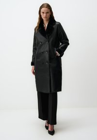 Manteau noir en simili cuir à double boutonnage avec un col en fourrure, poches latérales, porté par un mannequin vêtu de pantalons noirs et de talons.