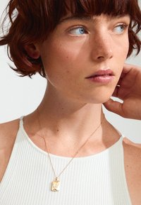 Collier en pendentif en or avec un charme rectangulaire, présentant un design floral, sur une fine chaîne, porté sur un haut blanc à côtes.