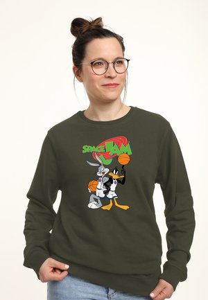 Kobieta w okularach, z włosami związanymi w kok, ubrana w oliwkowozieloną bluzę z nadrukiem postaci z bajek Bugs Bunny i Daffy Duck trzymających piłki do koszykówki oraz napisem "Space Jam".